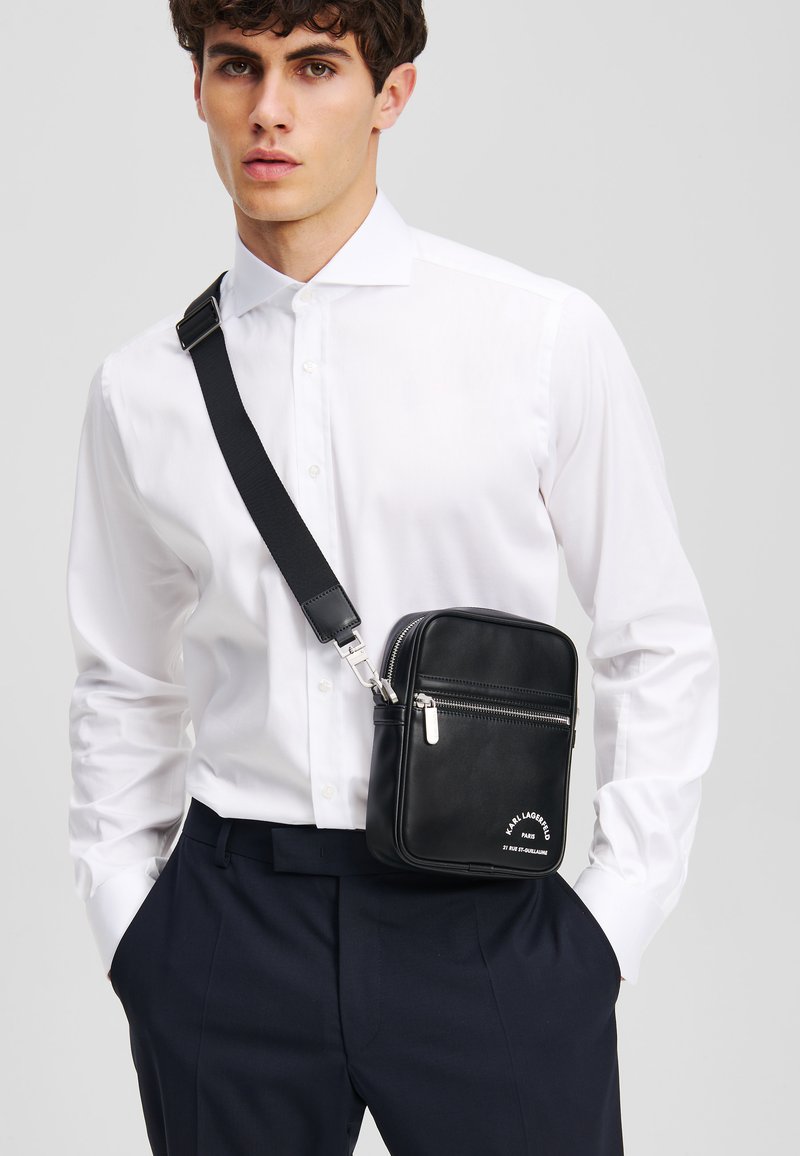 Čierna kožená crossbody taška so zipsovým uzáverom, nastaviteľným popruhom a embosovaným logom. Vyznačuje sa hladkou textúrou a kompaktným obdĺžnikovým tvarom.