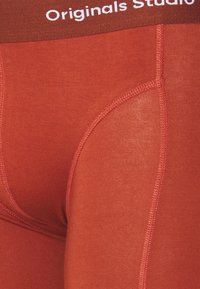 Rote Leggings mit glatter Textur, ausgestattet mit einem elastischen Bund, der mit "Originals Studio" beschriftet ist, und Kontrastnähten entlang der Nähte.