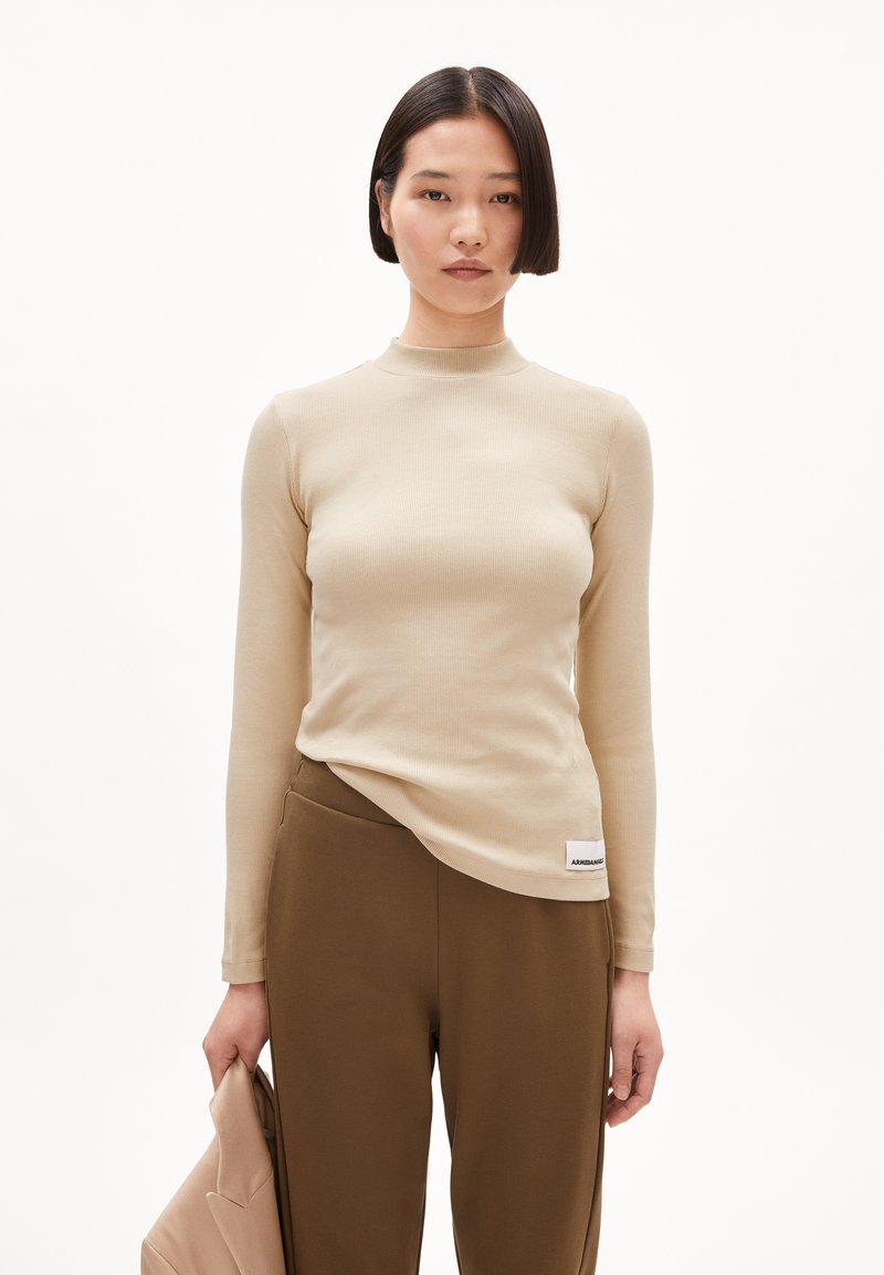Beige Langarmoberteil mit einem Stehkragen, geripptem Textur und figurnahem Design, kombiniert mit braunen Hosen und einer passenden Jacke.