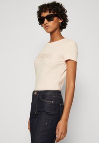 Camiseta ajustada de color rosa claro con un logotipo texturizado de "GUESS LOS ÁNGELES" en detalle metálico, combinada con jeans de mezclilla oscura con costuras doradas.