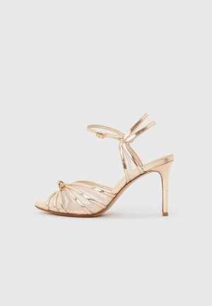 Goldfarbene, römische High-Heel-Sandale mit Knöchelschnalle, offener Spitze und Knoten-Detail vor hellem Hintergrund.