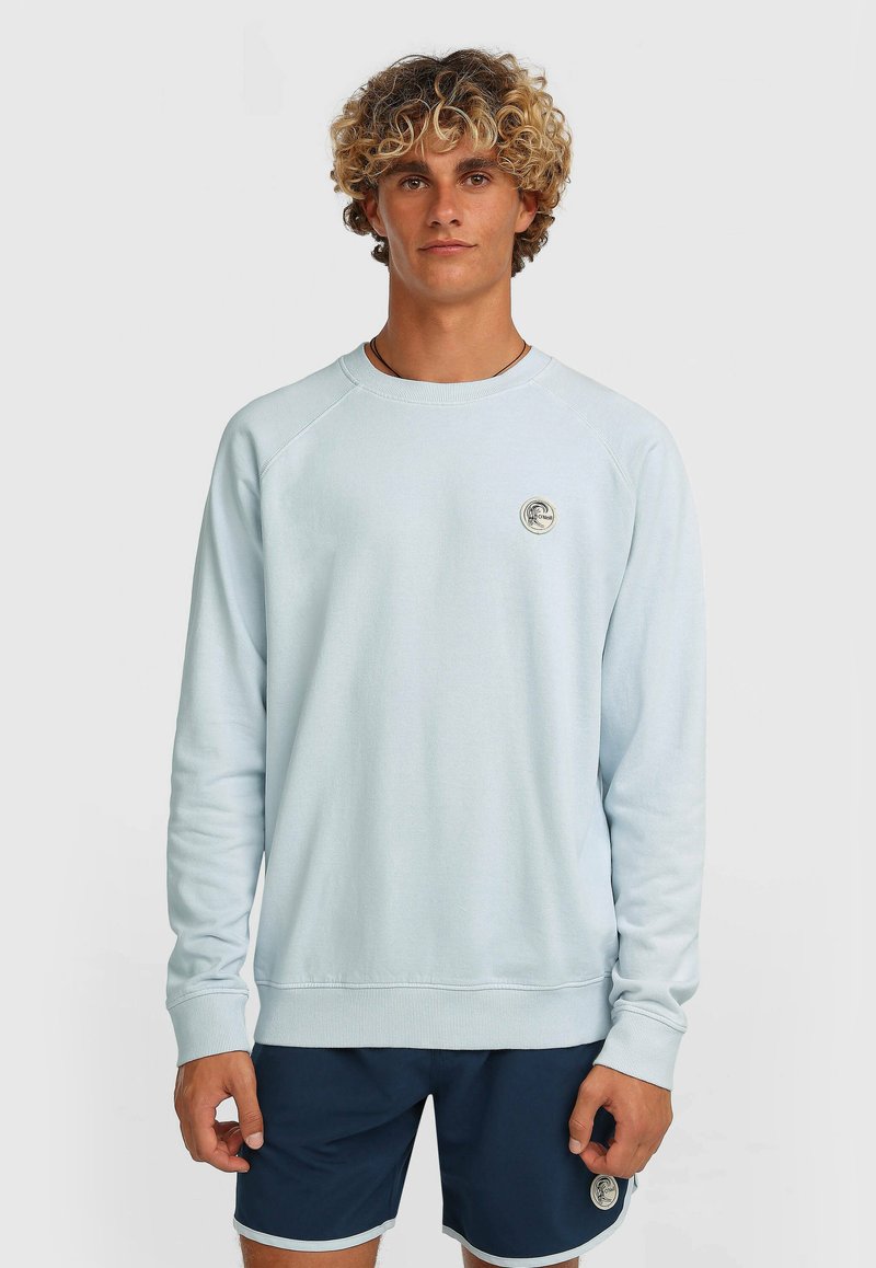 Sudadera azul claro con mangas largas raglán, puños y dobladillo acanalados. Presenta un parche de logo circular en el pecho. Tejido suave y con textura.