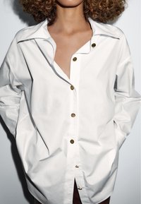 Bílá nadměrná košile z bavlny, s hlubokým výstřihem do V, velkým límcem a výraznými zlatými knoflíky na přední straně.