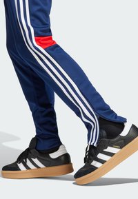 Pantalones deportivos de color marino con rayas blancas y acentos rojos; combinados con zapatillas negras y blancas con suela de goma y la marca "Samba".