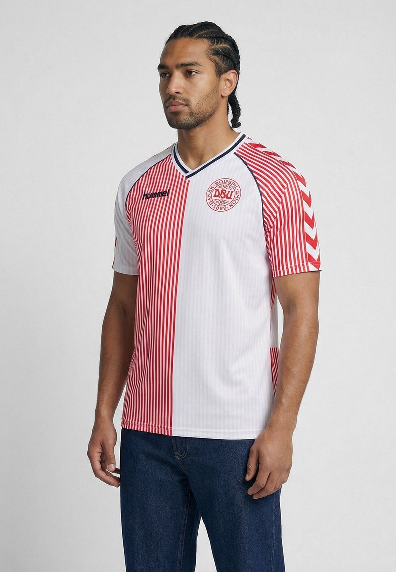 Homme aux cheveux tressés portant un maillot de football du Danemark rayé verticalement rouge et blanc et un jean bleu, tourné vers la gauche sur un fond uni.