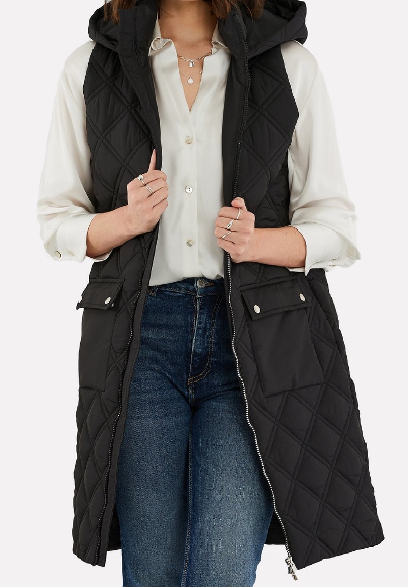 Femme portant un gilet matelassé noir sans manches avec capuche par-dessus un chemisier blanc à boutons et un jean bleu, tenant les bords du gilet avec les deux mains.