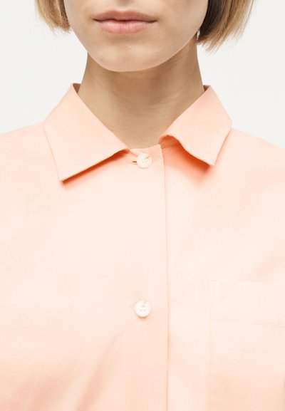 Κοντινή φωτογραφία ενός ατόμου που φορά ένα ελαφρύ ροδακινί πουκάμισο με κουμπιά, γιακά και τσέπη στο στήθος, εμφανίζοντας τον λαιμό και το κάτω μέρος του προσώπου.