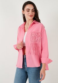 Light pink button-up shirt with a textured fabric, featuring a lace panel and fringe detail on the front pocket. Styled with blue jeans.