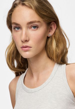 Portrait en gros plan d'une personne aux cheveux longs et ondulés. Elle porte un débardeur gris clair côtelé et de petites boucles d'oreilles décoratives.