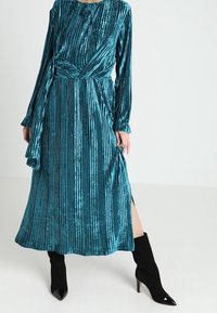 Tyrkysové sametové šaty s vertikálními pruhy, dlouhými rukávy a páskem v pase. Obsahují boční rozparek a jsou v kombinaci s černými vysokými podpatky.