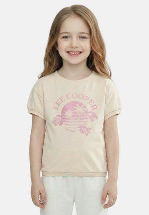 Jeune fille souriante aux longs cheveux châtains clairs, portant un T-shirt beige Lee Cooper avec un motif tropical rose et un pantalon blanc.