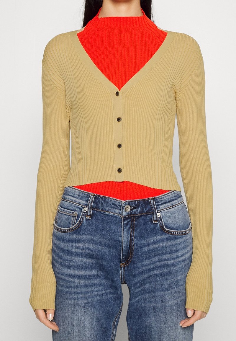 Cardigan beige côtelé à manches longues et col en V, avec des boutons foncés, porté sur un haut côtelé rouge vif et un jean bleu.