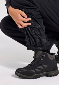 Schwarze Wanderschuhe mit einem strukturierten grauen Stoff, robusten Sohlen und gepolstertem Kragen. Schwarze wasserdichte Hosen mit elastischen Bündchen. Hand, die die Hosen anpasst.