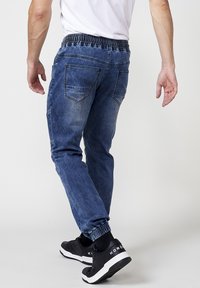 Pantalones jogger de mezclilla en azul oscuro con cinturilla elástica, tobillos acampanados y bolsillos traseros. Se llevan con zapatillas negras.
