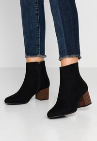 Botines de ante negros con punta afilada, cremallera lateral y tacón rectangular marrón texturizado. Combinados con jeans ajustados oscuros que tienen puños deshilachados.
