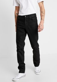 Hombre vistiendo jeans negros ajustados, zapatillas blancas y una camiseta blanca, apoyado contra un fondo liso claro que muestra tatuajes en ambos antebrazos.