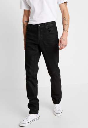 Man som bär snäva svarta jeans, vita sneakers och en vit t-shirt, står mot en enkel ljus bakgrund och visar tatueringar på båda underarmarna.