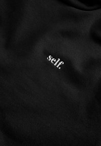 Tessuto di cotone nero con un piccolo ricamo bianco che dice "self." posizionato sul davanti, che mette in risalto una texture liscia.