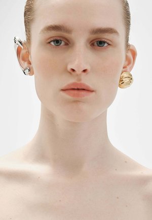 Primer plano de una persona con piel pálida y cabello peinado hacia atrás, que lleva pendientes metálicos desiguales: un audaz brazalete plateado a la izquierda y un aro dorado texturizado a la derecha.