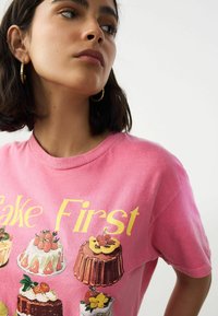 Pink cotton t-shirt featuring cake illustrations and the phrase "Bake First" in yellow. Round neckline, short sleeves, standard fit.