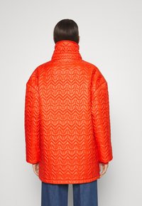 Czerwona krótka pikowana kurtka z wysokim kołnierzem, zygzakowatym wzorem i luźnym krojem. Rękawy szerokie, tkanina wydaje się ocieplana.