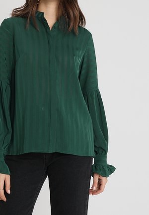 Kobieta ubrana w ciemnozieloną bluzkę z długim rękawem, z delikatnymi paskami i falbanianymi mankietami, połączoną z czarnymi spodniami na jednolitym tle.