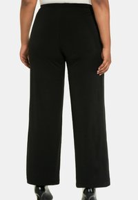 Osoba w czarnych szerokich spodniach i czarnych szpilkach, pokazana od pasa w dół, stojąca na jednolitym tle.