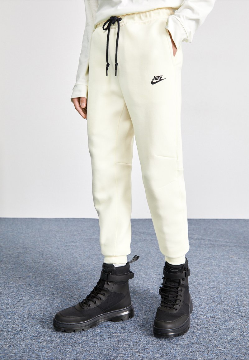 Weiße Jogginghosen mit elastischem Bund und schwarzem Kordelzug sowie Nike-Logo; kombiniert mit schwarzen, robusten High-Top-Stiefeln, die Schnürsenkel und einen Knöchelriemen haben.