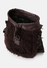 Saco têxtil castanho e peludo com fecho de cordão, detalhes em vinil e alças ajustáveis; apresenta um detalhe de marcação na lateral.
