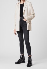 Osoba v krémovém blejzru, černém topu, tmavých skinny džínách a černých lesklých šněrovacích kotníkových botách stojící na bílé pozadí.