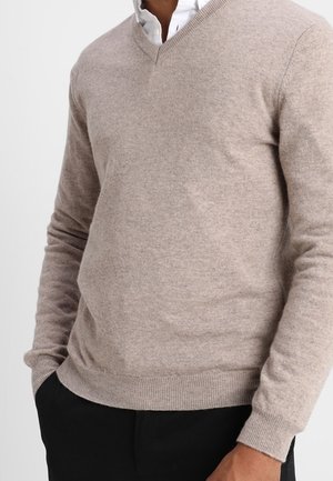 Mann trägt einen beigen V-Ausschnitt-Pullover über einem weißen Hemd mit Kragen und schwarzen Hosen, Hände teilweise in den Taschen, vor einfarbigem Hintergrund.