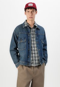 Young man wearing a red cap, denim jacket over a gray plaid shirt, and brown pants, standing with hands in pockets against white background.