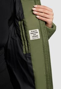 Indersiden af en grøn quiltet dynejakke med sort for, med en stofmærkat der lyder, "En varm kram lavet af stof."