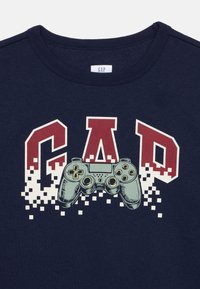 Μπλε σκούρο βαμβακερό φούτερ με την επιγραφή "GAP" σε κόκκινα και κρεμ γράμματα, με γραφικό pixel ενός γκρι χειριστηρίου βιντεοπαιχνιδιών.