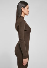 Brun ribbet langermet topp med høy hals, tettsittende design og teksturert stoff, vist på en modell fra en sidevinkel.