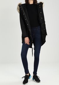 Svart parka-jacka med huva i konstpäls, med detaljer i guld, kviltad textur, i kombination med mörkblå slim-fit-jeans och svarta sneakers.