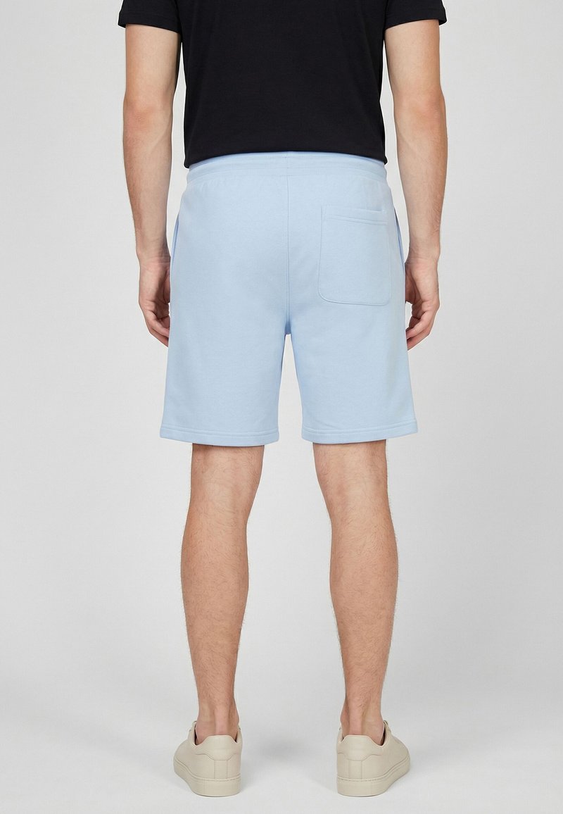Homme portant un short bleu clair et des baskets beige, debout de dos devant un fond uni.