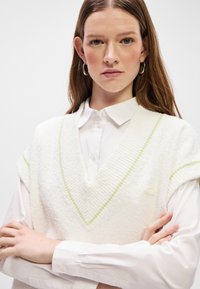 Λευκό πλεκτό γιλέκο με V λαιμόκοψη και κοντά μανίκια, με πράσινη διακόσμηση στις άκρες, φορεμένο πάνω από ένα λευκό πουκάμισο με γιακά.