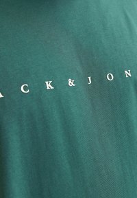 Donkergroene t-shirt met witte bedrukking met de tekst "ACK & JON" gecentreerd op de voorkant. Glad materiaal en klassiek ontwerp met ronde hals.