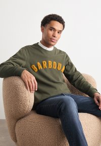 Olivově zelená mikina s nápisem "BARBOUR" v žlutých texturovaných písmenech, nošena přes bílou košilovou košili. Model sedí na béžovém huňatém křesle.