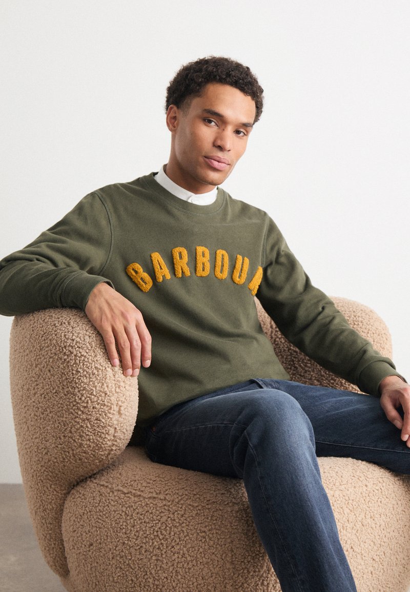 Olivově zelená mikina s nápisem "BARBOUR" v žlutých texturovaných písmenech, nošena přes bílou košilovou košili. Model sedí na béžovém huňatém křesle.