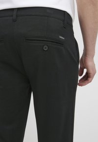 Schwarze Hose aus strukturiertem Stoff, mit zwei Gesäßtaschen und Knopfverschluss sowie einem aufgenähten Etikett am Bund.