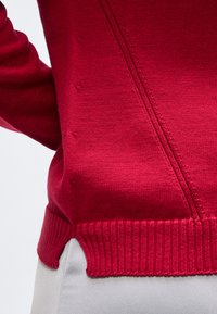 Κοντινό πλάνο σε κόκκινο πλεκτό πουλόβερ με πλαϊνή σχισμή, διαγώνια λεπτομέρεια ραφής και ελαστική μέση, φορεμένο πάνω από ανοιχτό γκρι παντελόνι.