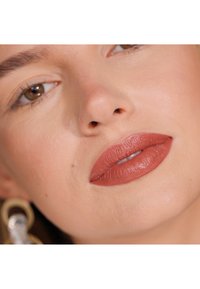 Matte terracotta lipstick on full lips, smooth texture, natural skin tone, with subtle shine. Background features blurred earrings.
