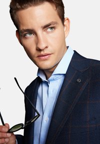 Granatowa marynarka w kratę z teksturowanym wykończeniem, jasnoniebieska koszula z guzikami oraz czarne okulary przeciwsłoneczne trzymane w ręku. Wyjątkowe detale to logo na marynarce.