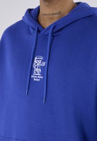 Blaues Hoodie mit einer Cartoon-Pizzastückfigur, die zeigt, und dem Text "Slice, Slice Baby!", der auf der Brust zu sehen ist. Das Nackentattoo der Person trägt die Aufschrift "Glaube an dich selbst."