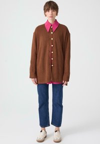 Brown ribbed oversized cardigan with a pink collar and large buttons, paired with blue straight-leg jeans and white sneakers.