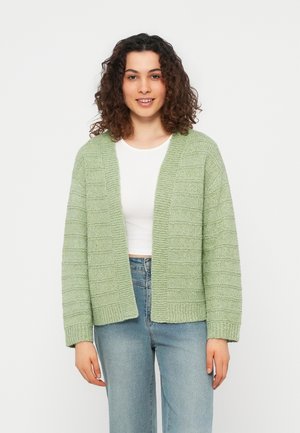 Femme aux cheveux bouclés portant un cardigan tricoté vert clair, un crop top blanc et un jean bleu, debout devant un fond uni.