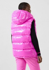 Gilet imbottito rosa con una texture lucida e trapuntata e cappuccio. Presenta un design ampio senza maniche, abbinato a pantaloni rosa aderenti.
