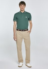 Zelená polo trička s krátkým rukávem, s bílozelenými pruhy, spárované s béžovými kalhotami a bílými teniskami. Hladký materiál, klasický střih.